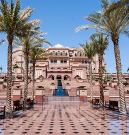 The image shows a low angle view of the entrance to a luxurious palace in Dubai. The entrance is surrounded by palm trees. Dubai is a city of luxury and excess, and this palace is a perfect example of that.