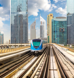 A Dubai Metro train is speeding towards the viewer, its headlights shining brightly. The railings of the track can be seen, and the tall buildings of Dubai are in the blurry background. The image shows one of the many ways to get around Dubai, and it highlights the city's commitment to public transportation.