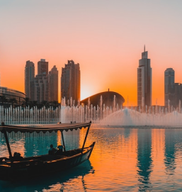 The Dubai Mall water fountain at sunset, with the city's skyline in the background. A boat passes in front of the fountain.